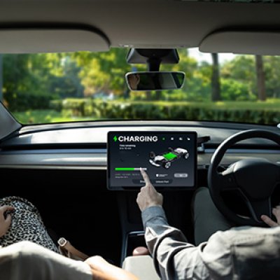 A photo taken inside an EV showing a driver and passenger in the front seat, the driver is pointing to the vehicle's charging screen.